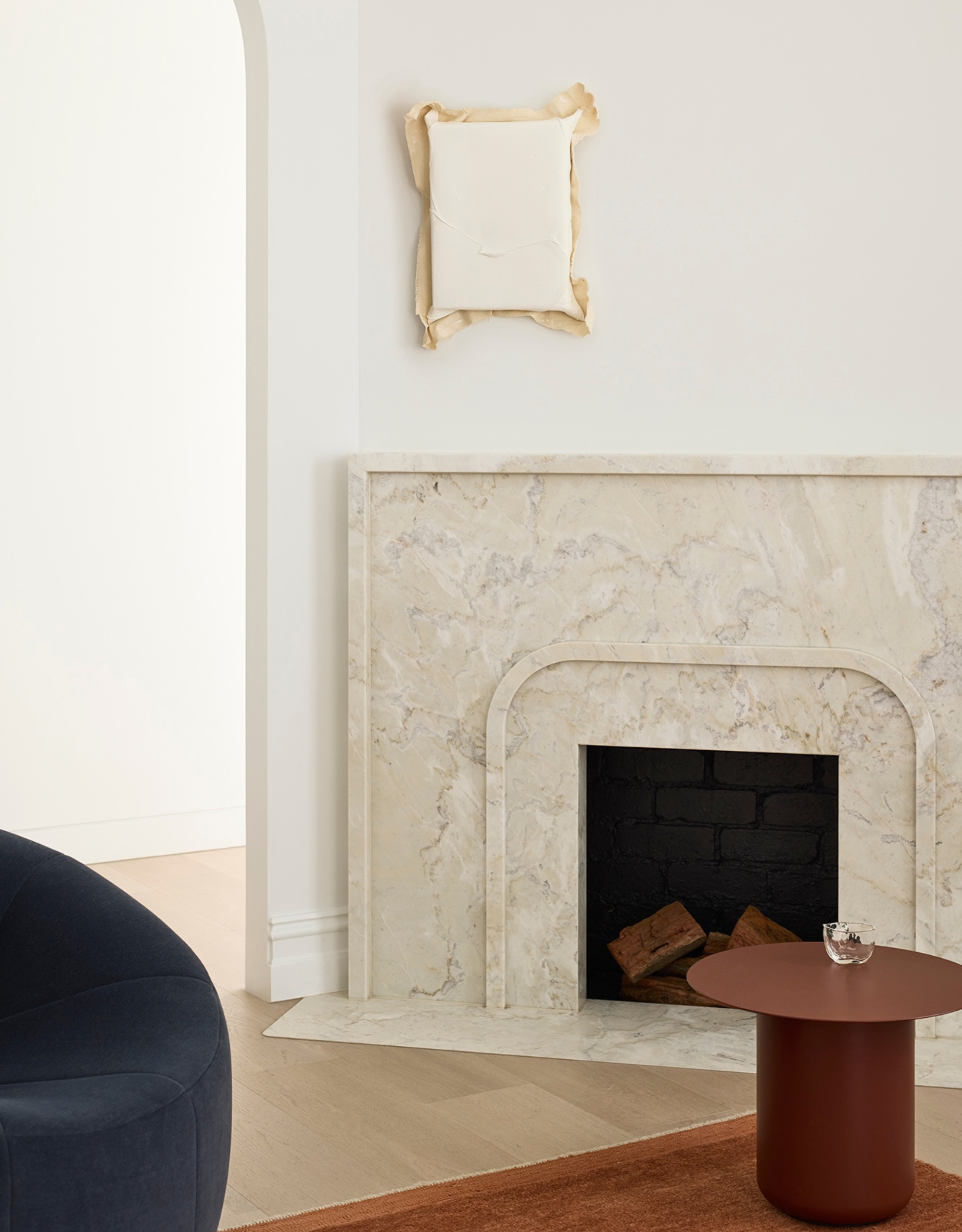 A white living room with a marble fireplace, a rust side table with a glass, a rust rug, and a blue chair