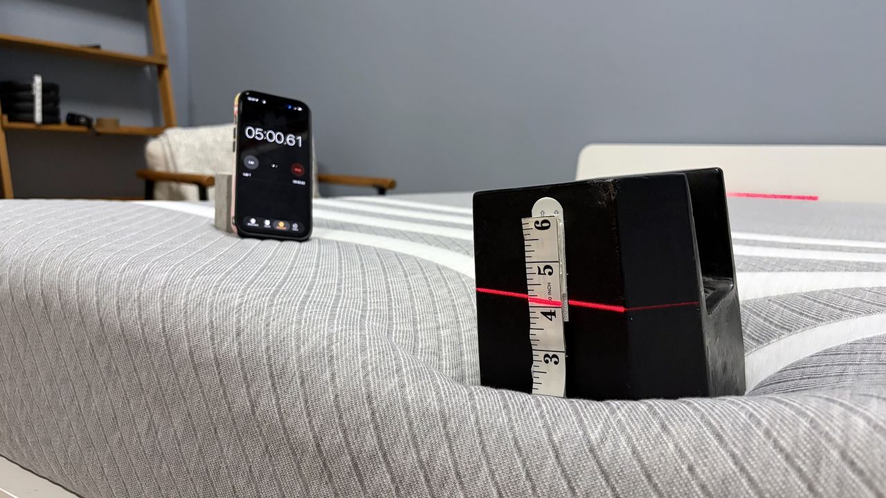 A close up of a 25kg weight balancing on the end of the Leesa mattress, a red laser line cutting through it indicates how far the weight has sank. To the left is a phone showing a five minute timer