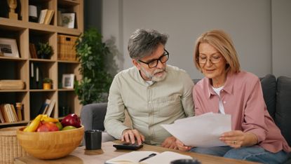 An older couple work on financial paperwork at their kitchen table.