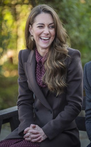 Kate Middleton wearing a long brown coat and burgundy polka dot dress sitting on a bench and laughing with green trees in the background