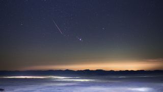 A meteor shower is pictured against a starry sky and picturesque foreground