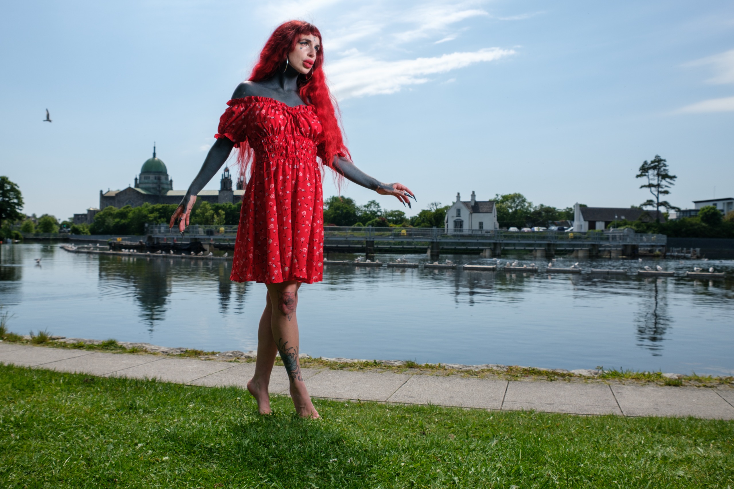 Red-haired person with dark body paint posing with both arms raised in a grassy field under a clear blue sky.