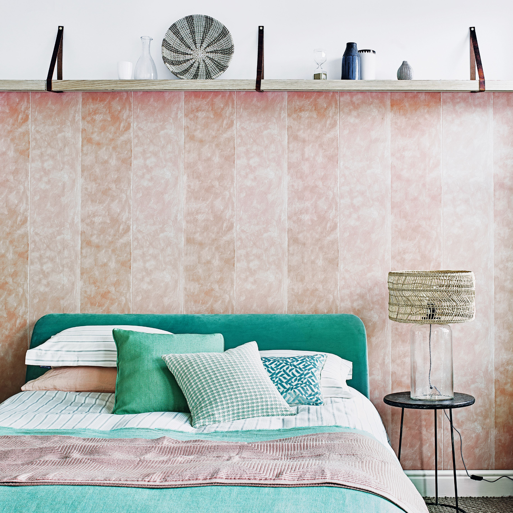 Bedroom with oak shelf hung at picture rail height on leather straps, over a double bed with green headboard with blush striped wallpaper