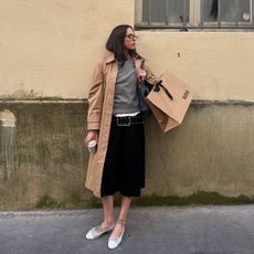 A woman wearing a tan trench coat, black culottes, white flats, and a gray sweater