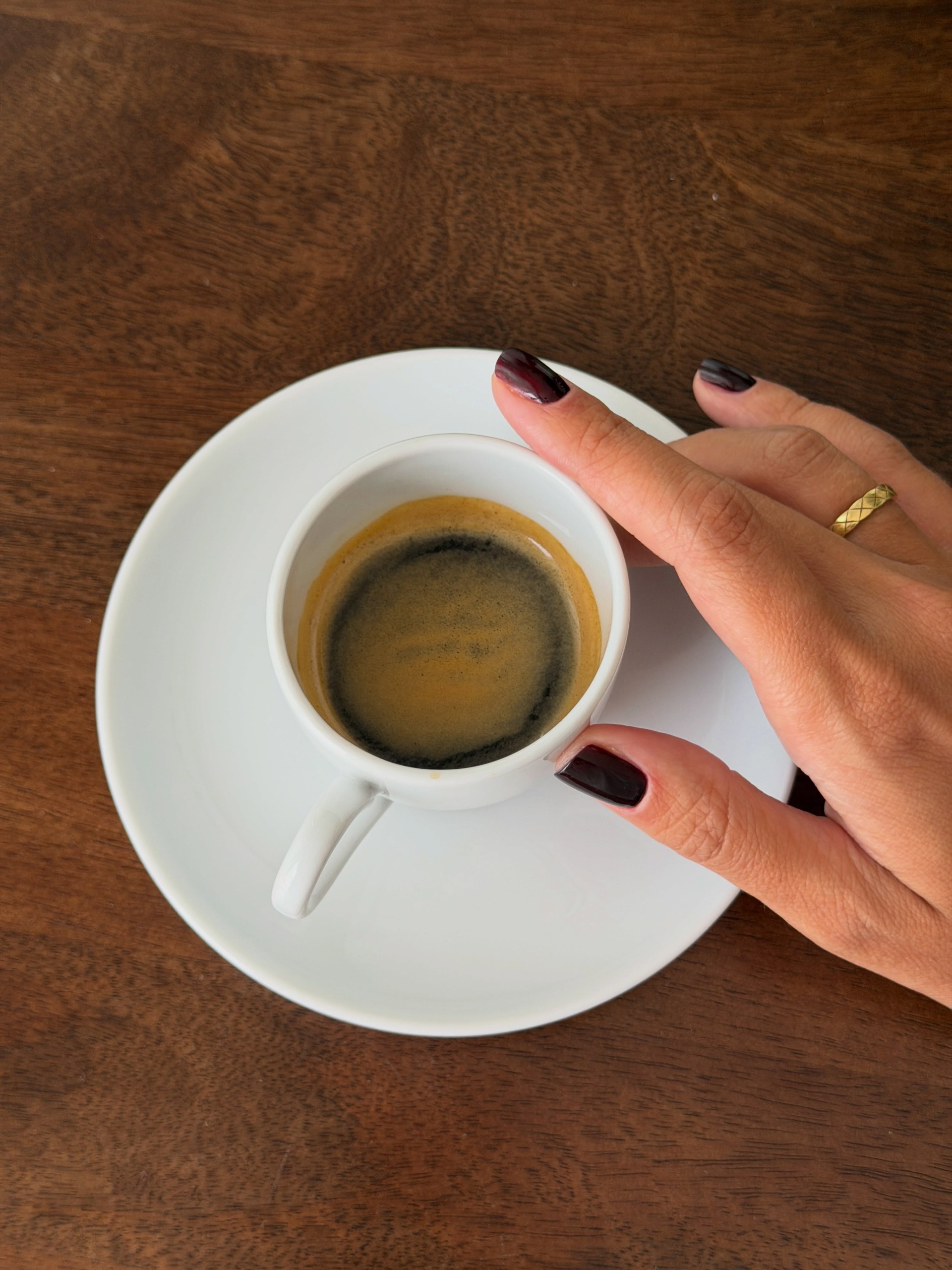 A hand with dark coffee nails reaches for a small cup of espresso on a white saucer sitting on a wooden table