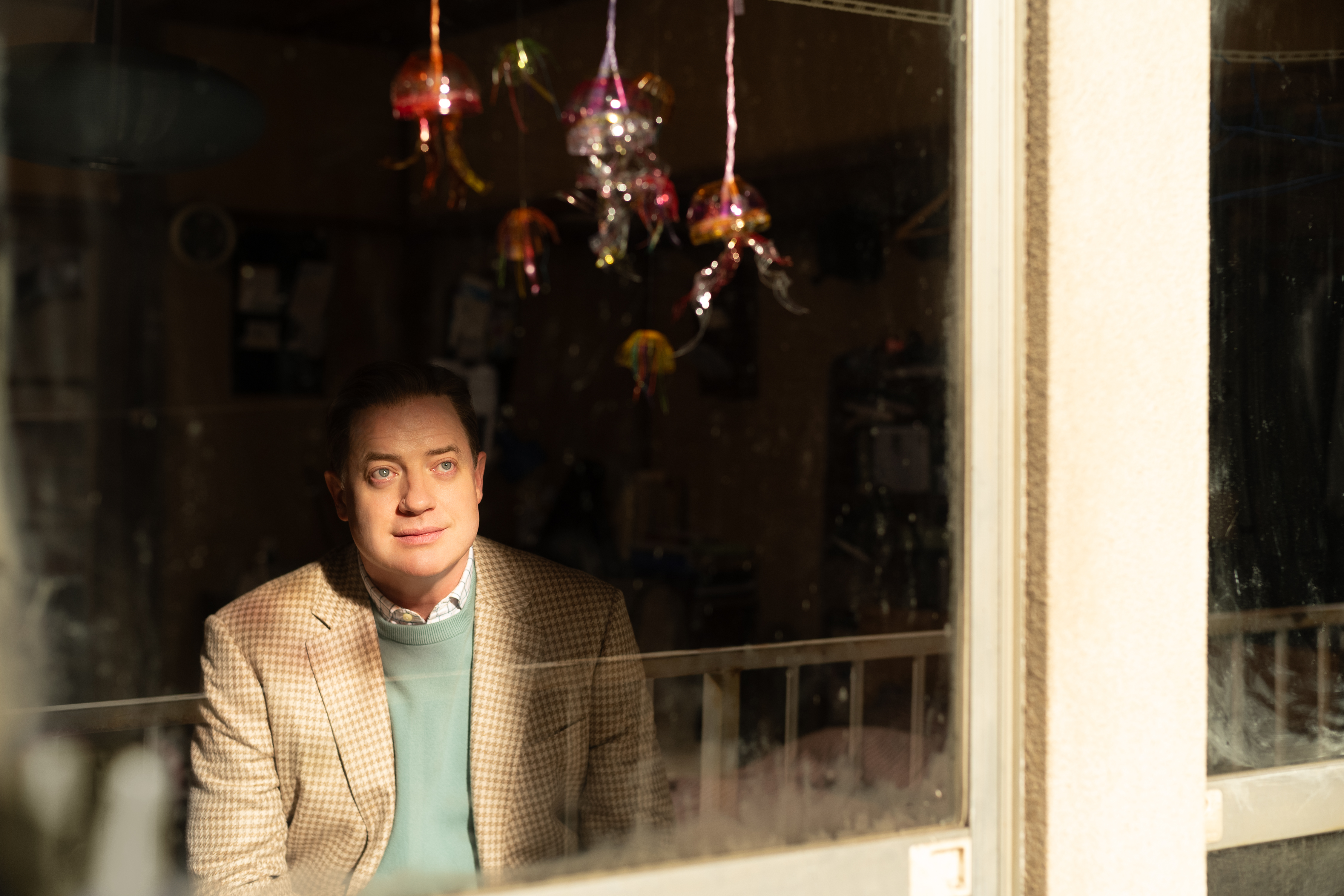 A white man (Brendan Fraser as Phillip Vandarploeug) sitting in a dark room, seen through a window with a white frame. He looks up at a child's art project sparkling in the sunlight. A film still from 'Rental Family.'