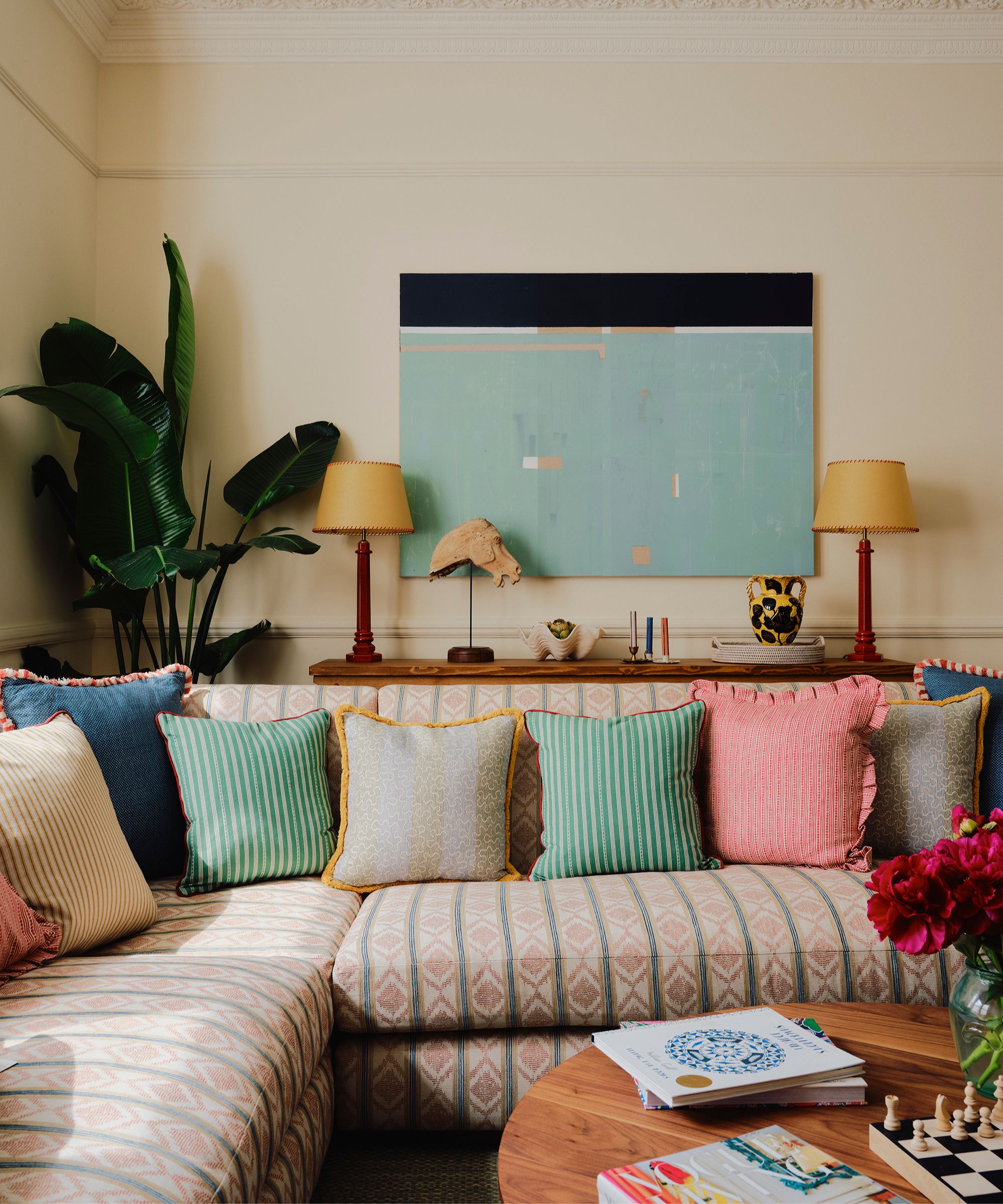 Living room with a patterned sectional sofa, colourful cushions, a console with two lamps on either side and antique artefacts in the middle, and a hidden TV mounted behind a canvas on the wall