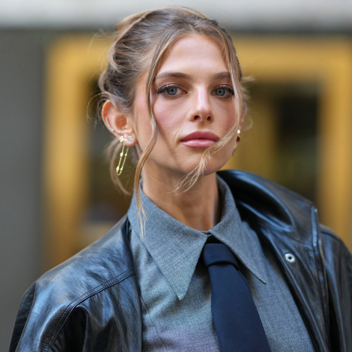 NEW YORK, NEW YORK - SEPTEMBER 15: A guest wears brown hair pulled back into a loose topknot with face-framing tendrils, small gold hoop earrings and multiple gold rings, an oversized black leather jacket, a grey button-down shirt with a pointed collar and a navy tie, outside TWP, during New York Fashion Week, on September 15, 2025 in New York, New York (Photo by Edward Berthelot/Getty Images)