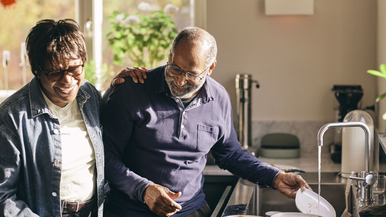 An older couple laugh as they wash dishes together in their kitchen.