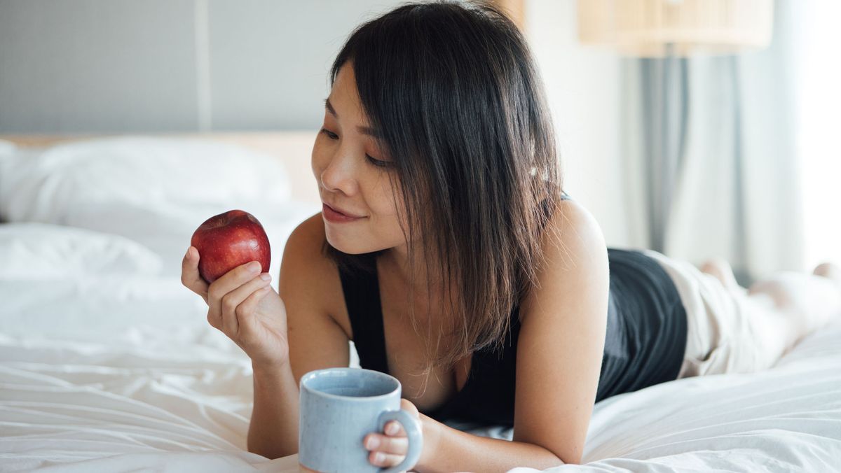 A woman with long dark hair lies in bed holding a red apple in one hand a cup of coffee in the other