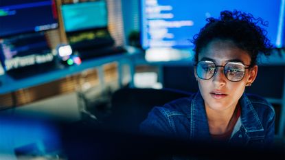 Female software developer using AI coding tools in a dimly lit office space, with desktop monitor reflecting in glasses and laptop devices in background.