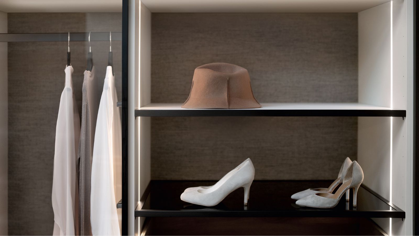 A modern wardrobe with built-in vertical LED lighting that softly illuminates the shelves and hanging space. On the shelves, a brown hat is displayed above a pair of elegant white high-heeled shoes, while a few light garments hang neatly to the side.