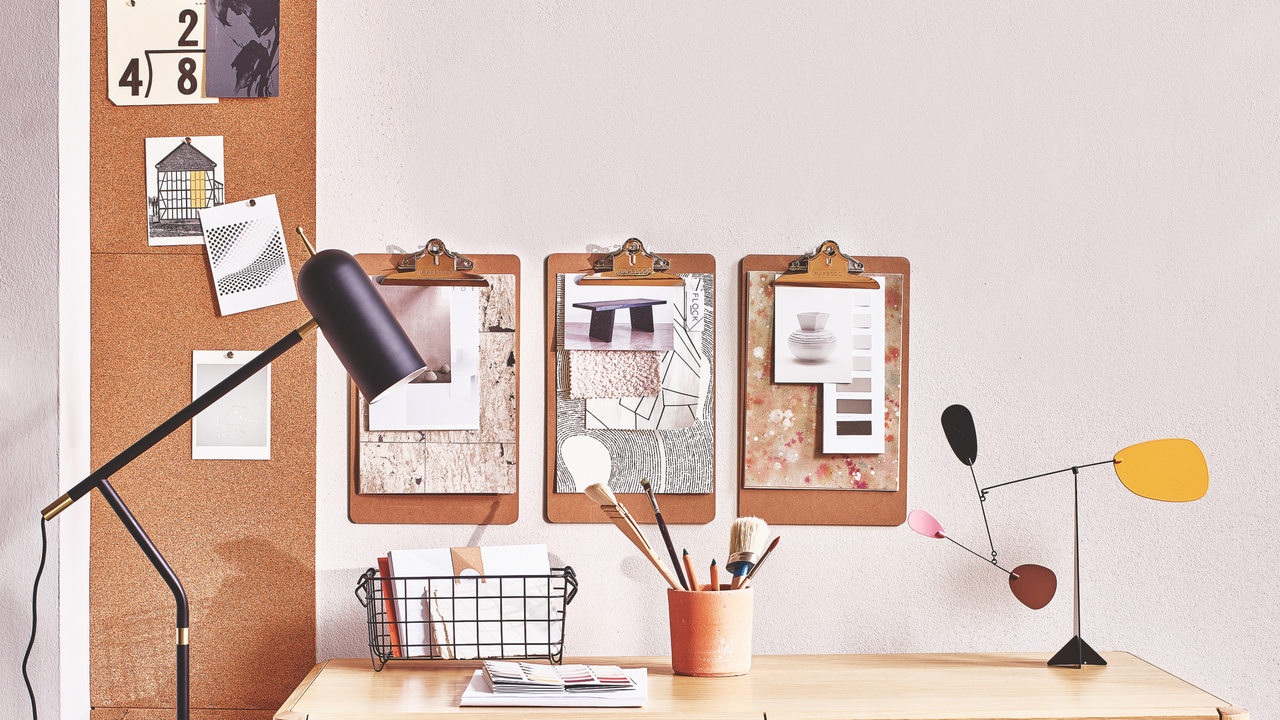 A wooden desk top with stationery and swatches of fabrics. There are three moodboards on the wall.