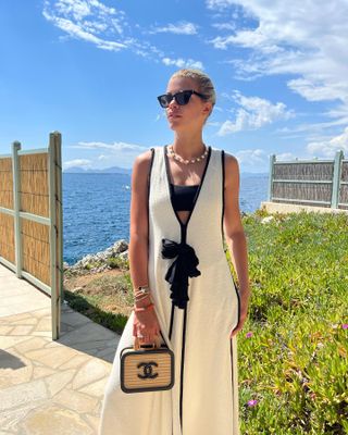 Sofia Richie Grainge standing near water wearing a white linen dress, a black strapless bikini top, and a string of pearls.
