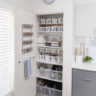Orgnaized pantry interior with categorized white boxes of foodstuffs, and hanging spice rack and towel rail on door back.