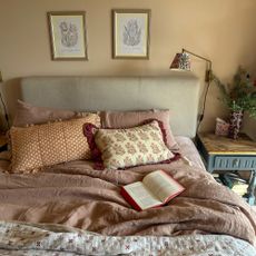Bed in neutral bedroom, with dusky pink linens and patterned cushions, beneath two framed illustrations