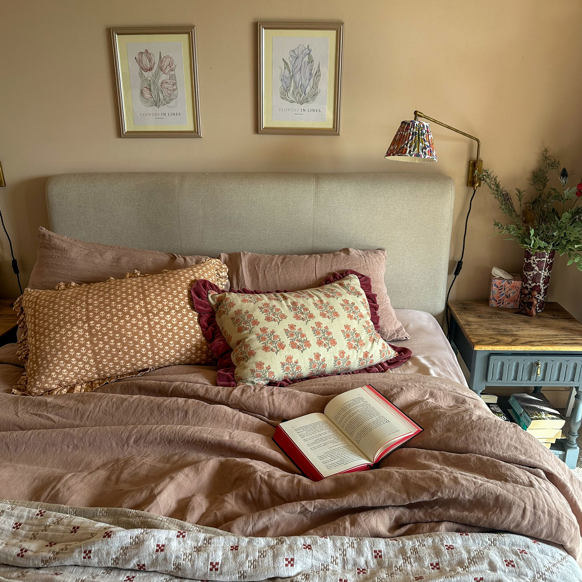 Bed in neutral bedroom, with dusky pink linens and patterned cushions, beneath two framed illustrations