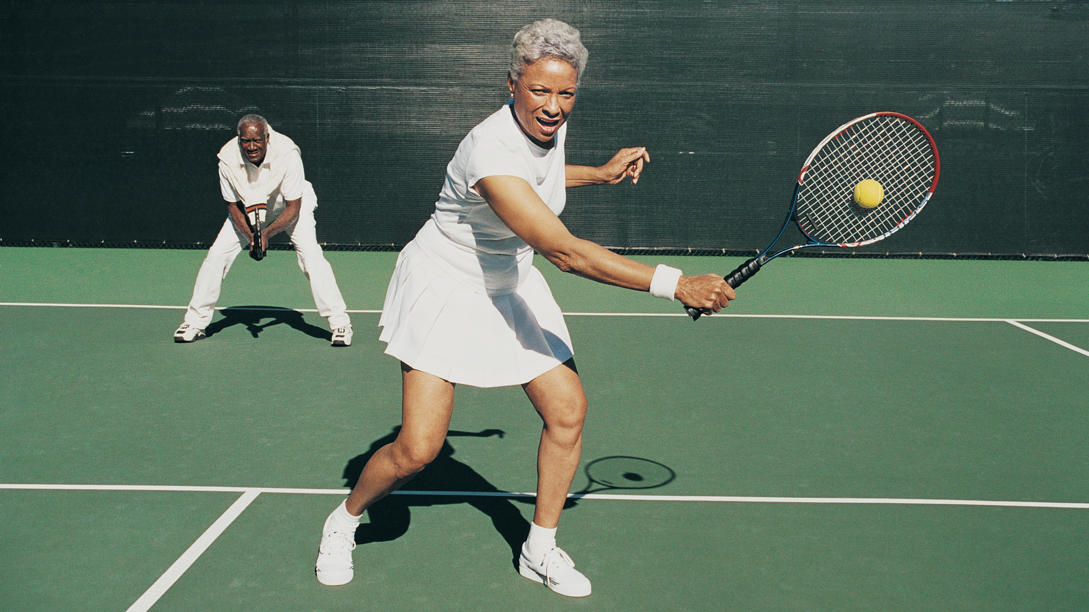 Woman striking a tennis ball with a man on the court behind her