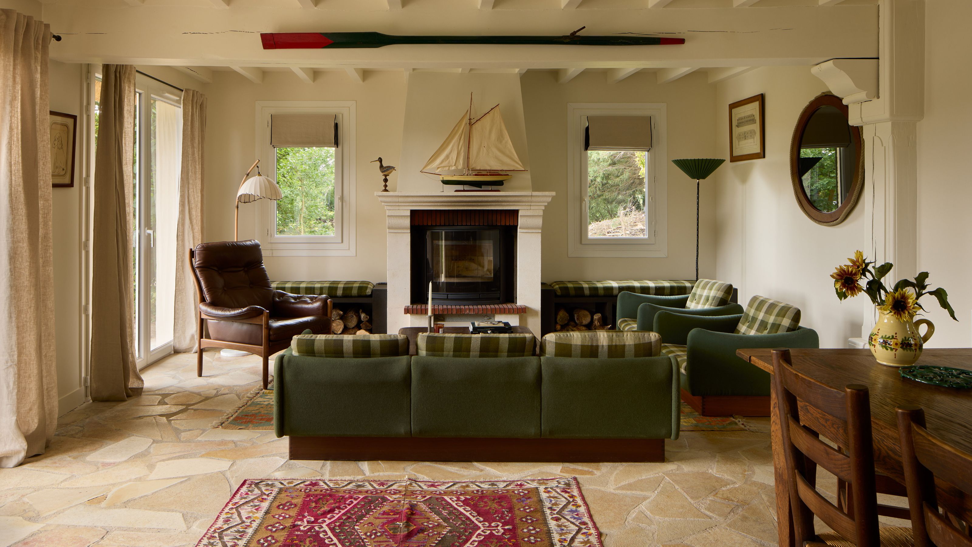 an open-plan living dining area in a french cottage in Normandy with lake and nautical nods like a vintage ship on the fireplace, green and plaid sofas and vintage wooden furniture