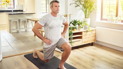 A man performs lunges at homes on an exercise mat with his hands on his hips. Behind him we see leafy plants, a breakfast bar and high stools.