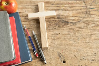 Wood Christian cross laying on a vintage wood desk with a stack of grey, red and navy blue books, a red apple and a pen and pencil
