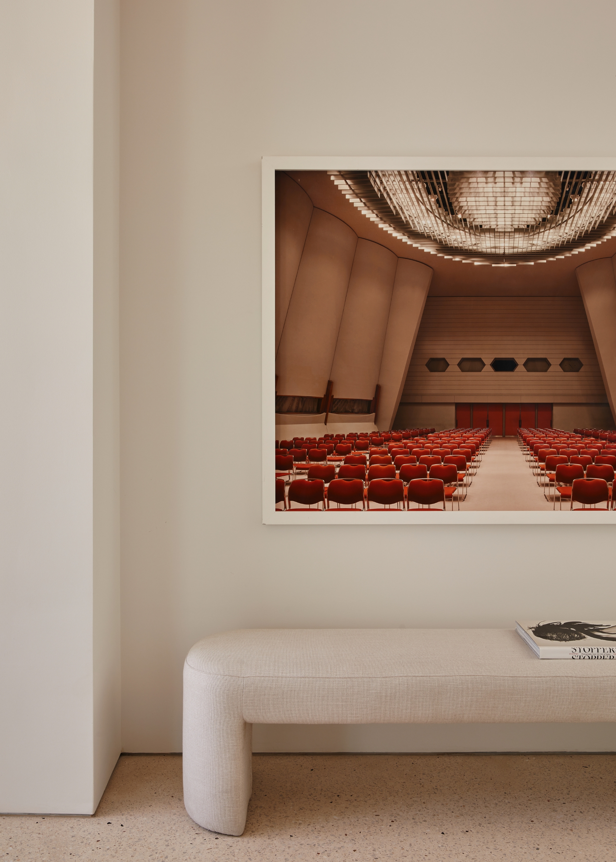 A white living room with a white bench, a coffee table book, and a framed photo of a room with red seats and a disco ceiling on the wall