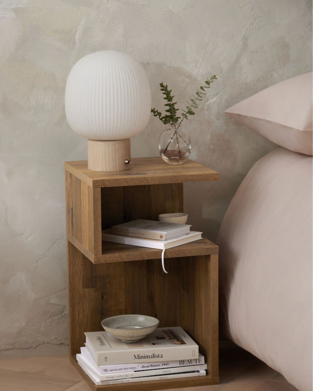 A wood bedside table with a zig-zag structure and built-in shelving, topped by an oval shaped table lamp with an opaque glass top and a wood base, a flower vase, books, and ceramics.