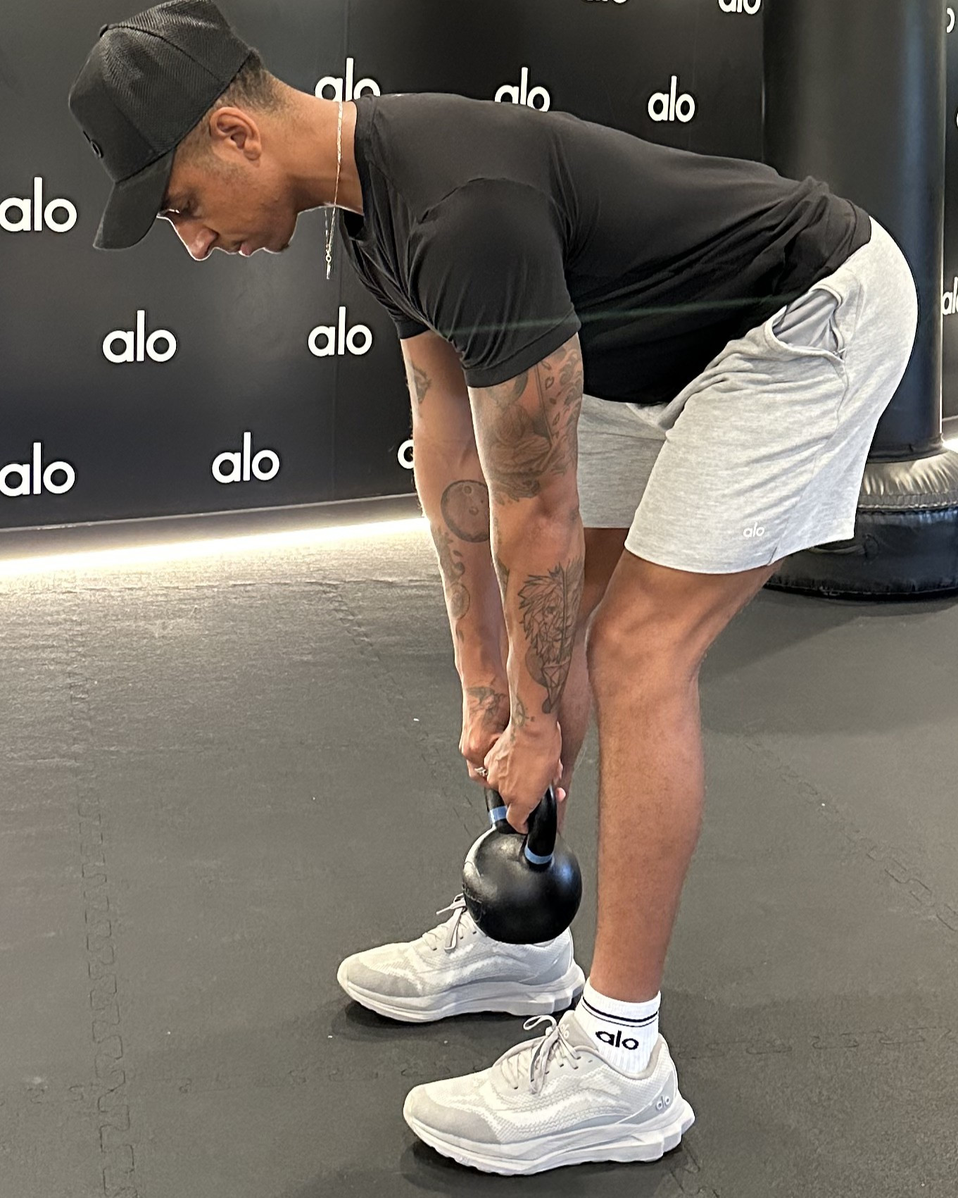 Man in black T-shirt, black cap, white shorts and white trainers demonstrates exercise