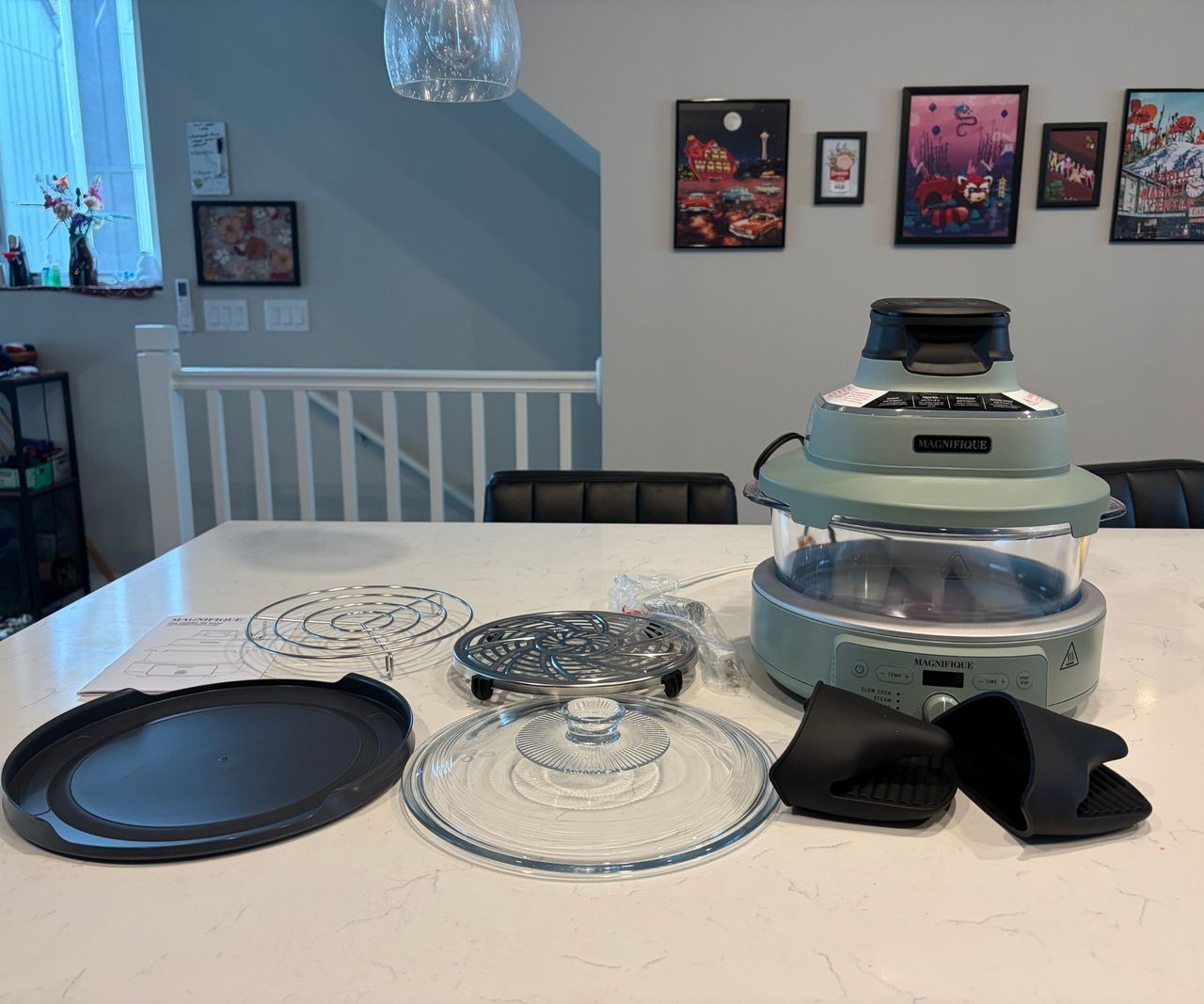 A Magnifique glass air fryer with a green base and top, on a white counter beside two wire trays, a black lid, a glass lid, and some siligone hand grips.