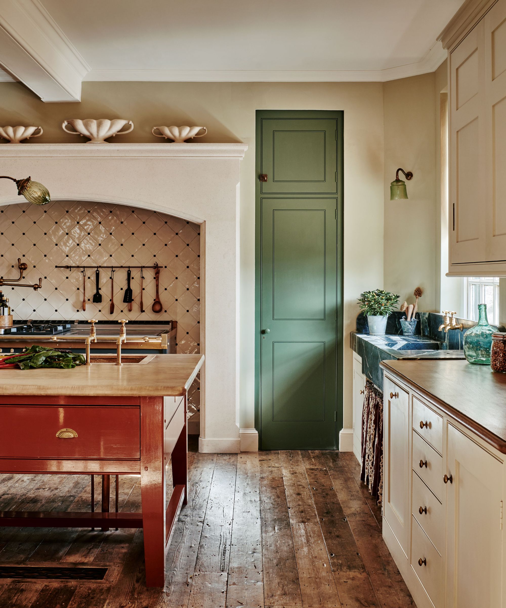 A traditional kitchen with cream walls and cabinets, a navy blue range cooker, a terracotta red freestanding island, wooden countertops, and a green-painted door to the pantry
