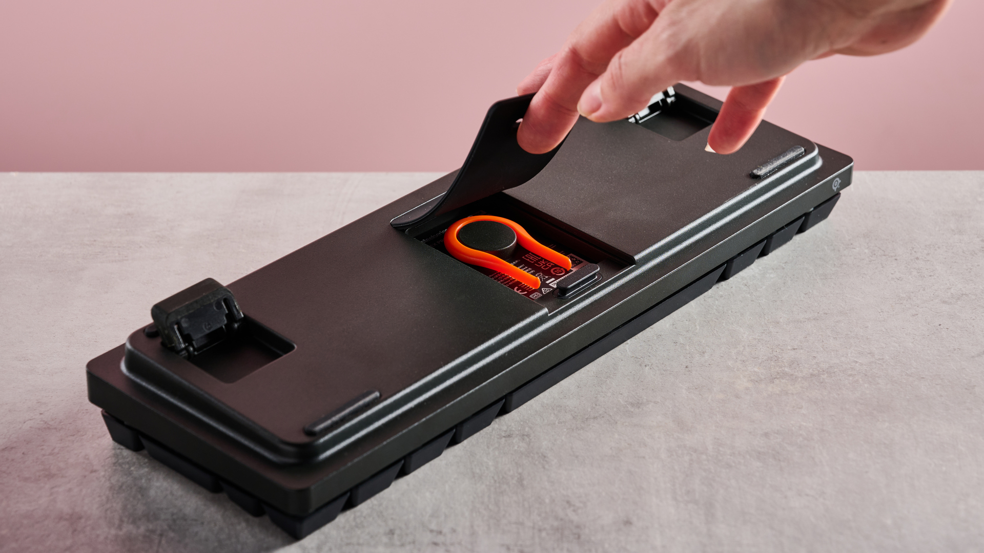 Underneath of SteelSeries Apex Pro Mini with hand holding keycap puller compartment flap, on desk with pink background