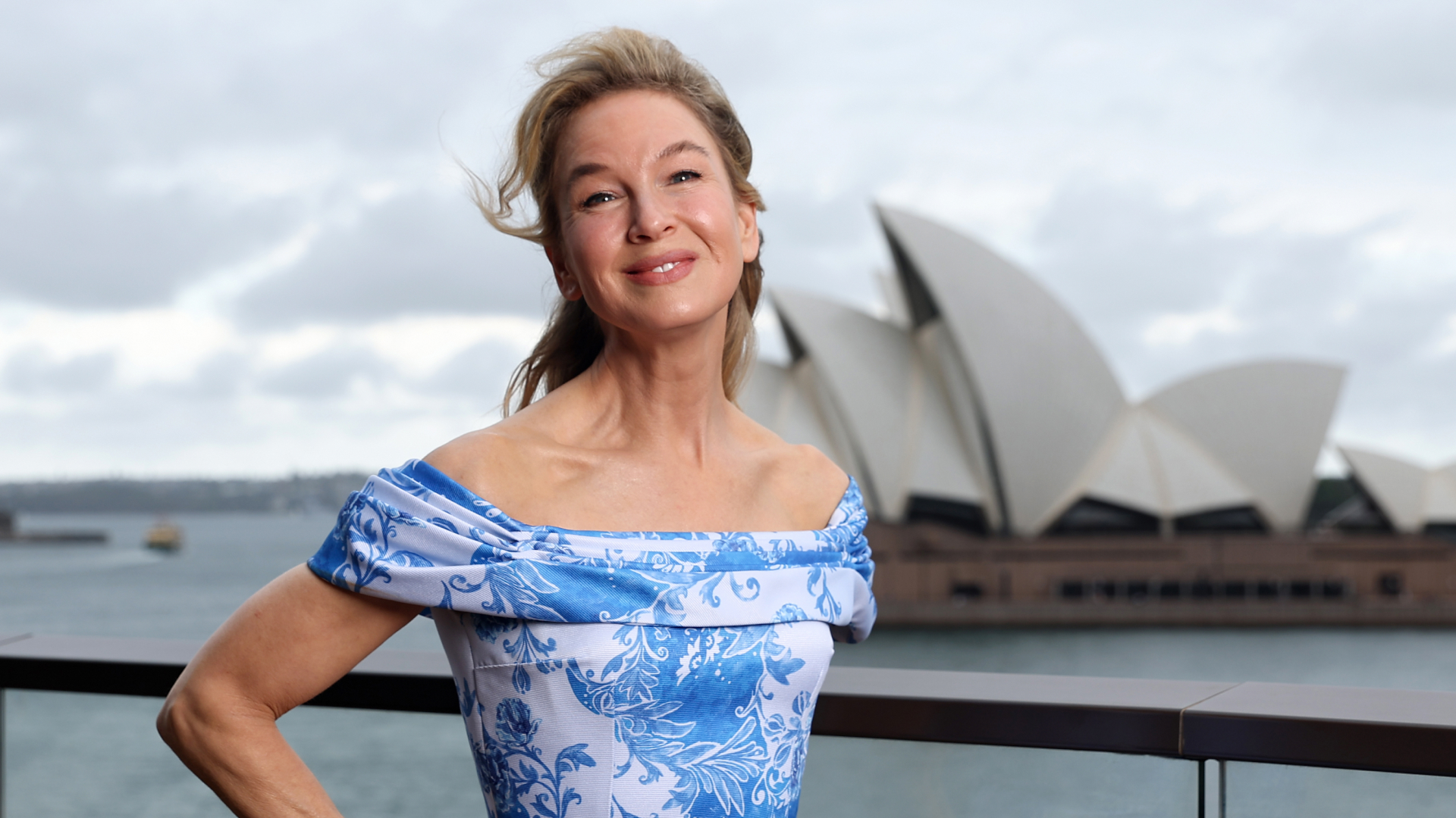 Renee Zellweger wearing an off-the-shoulder blue and white dress. She also is wearing a half up half down hairstyle and smiling at the camera with her hand on her hip.