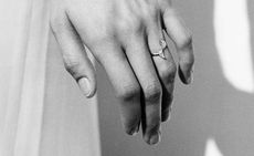 A black and white image focussing on a left hand with a ring the fourth finger. The ring has upward and downward triangle shape stones.