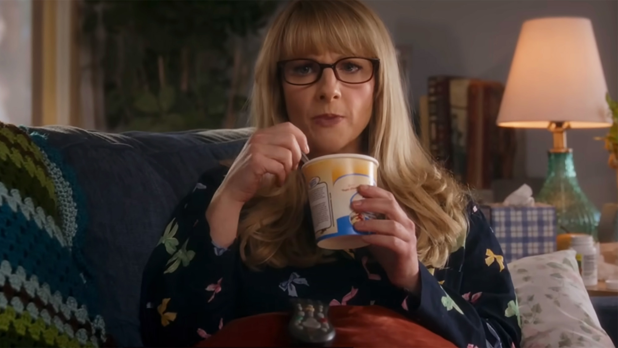 Melissa Rauch in her Bernadette glasses sits on a couch and eats from a pint on Stuart Fails to Save The Universe.