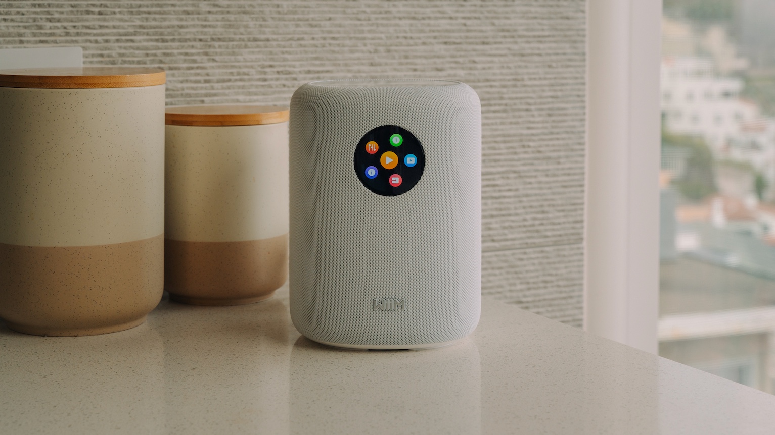 A white WiiM Sound smart speaker on a white surface in front of some jars.