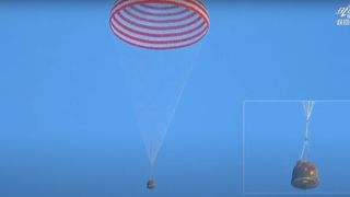 a dark-brown capsule descends through a blue sky beneath a red and white striped parachute.
