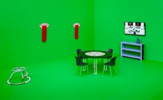 A dining area featuring circular dark green table and chairs and blue bookcase against a bright green wall and floor. A widescreen monitor above the bookcase displays a digital image of the dining area.