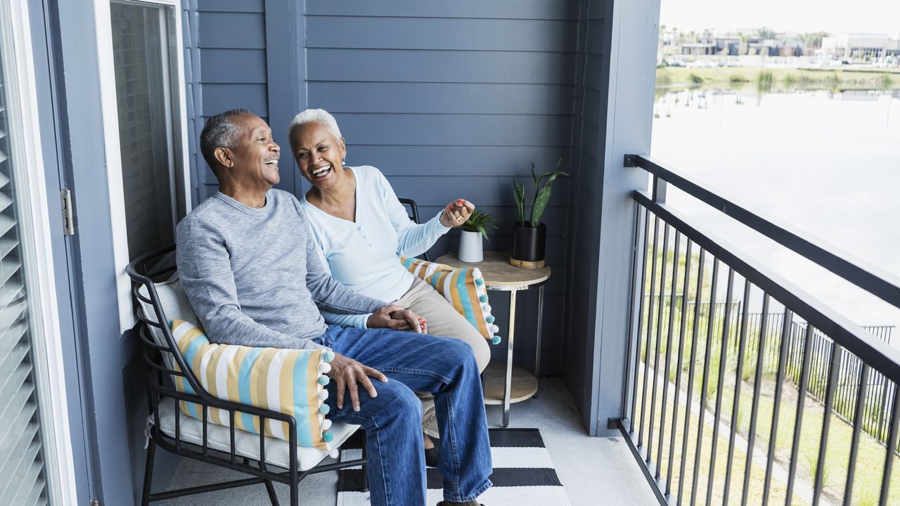 An older couple smile and laugh while sitting on the balcony of their apartment.