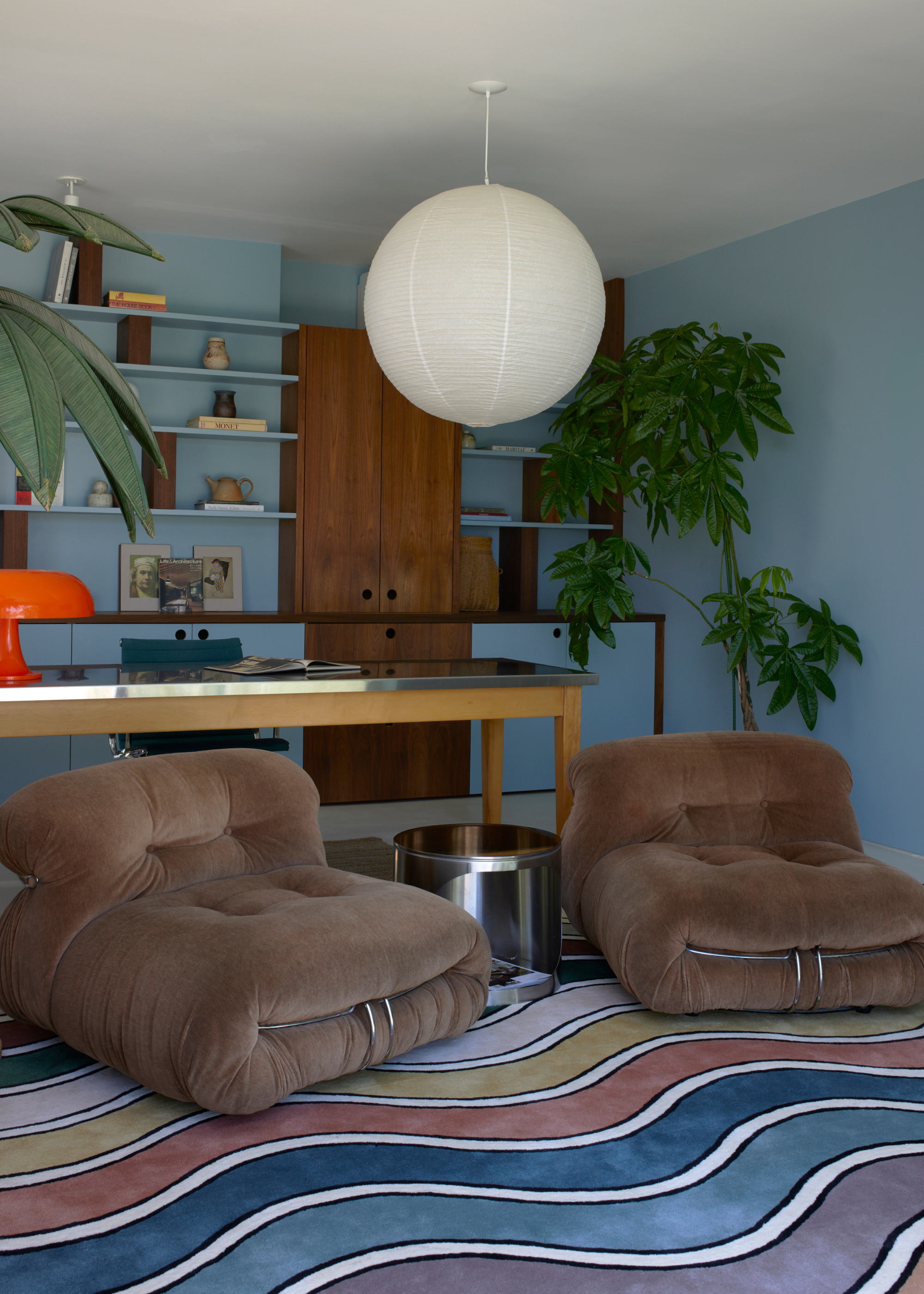 A blue room with a rice paper pendant, a potted plant, floating shelved, a desk, a brown sofa chair with a chrome frame