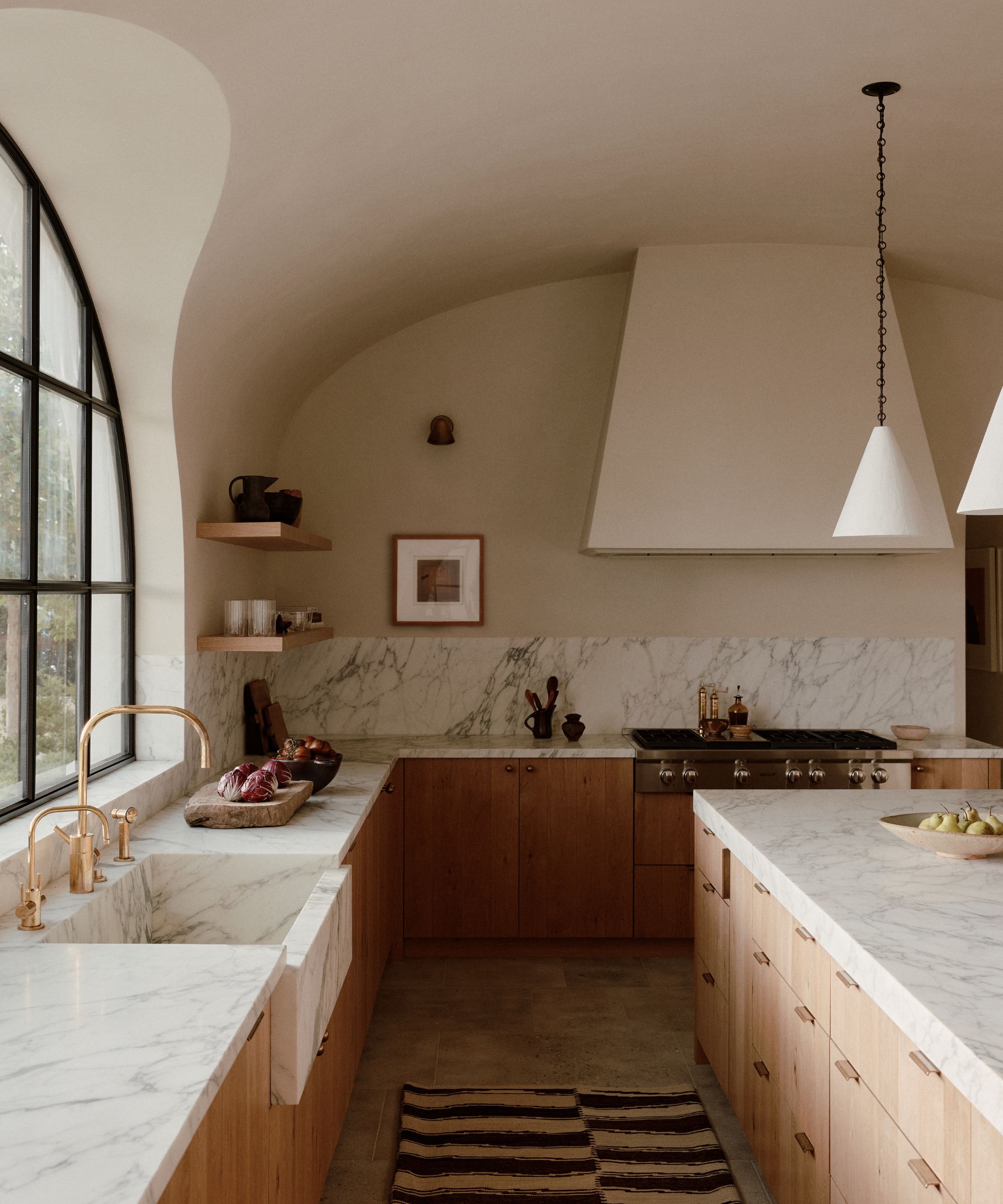 A detailed view of the kitchen workspace featuring a heavy marble farmhouse sink, unlacquered brass hardware, and a integrated marble backsplash. The scene includes a gas range under a minimalist, plastered vent hood