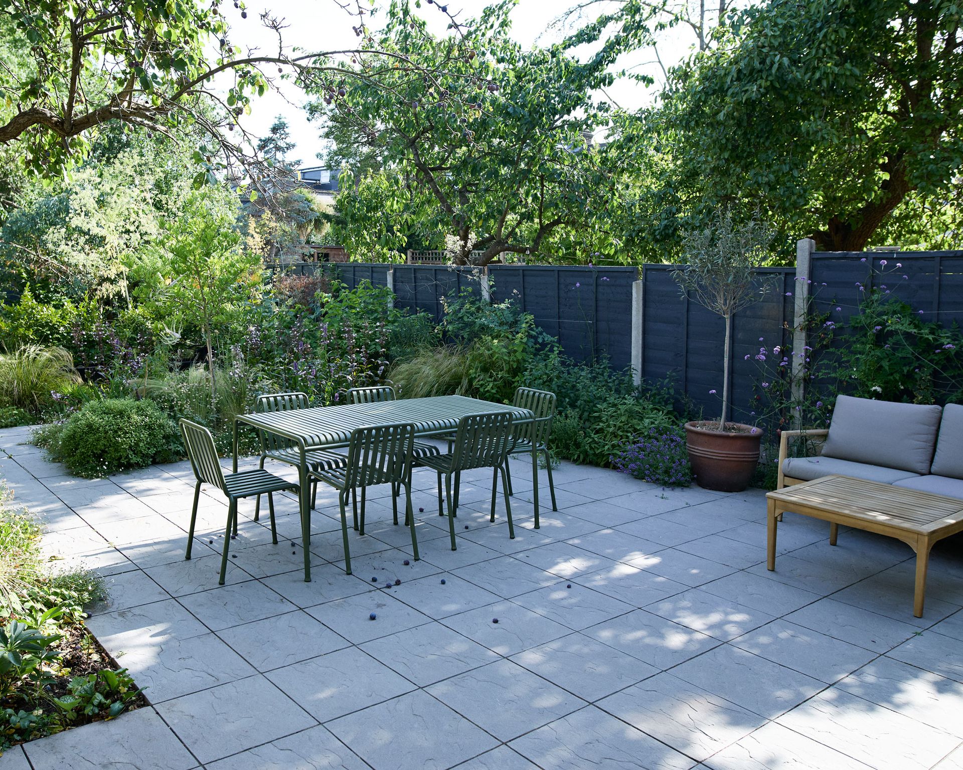 shady garden design with pale colored paving, black fence, outdoor furniture set, trees and shrubs
