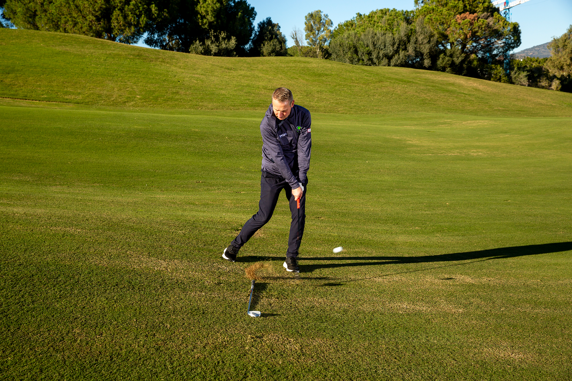 Anders Mankert demonstrating how to hit a pitch shot from a downhill lie with the correct ball position identified by a club on the ground