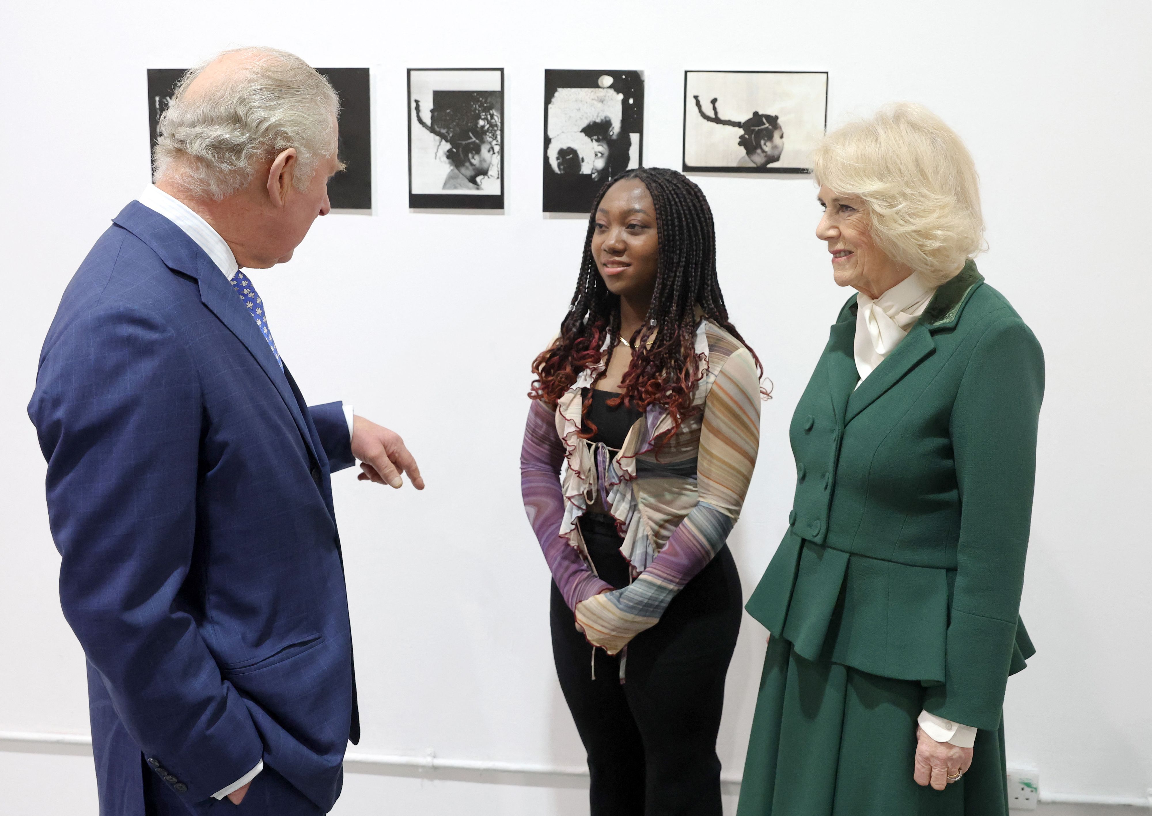 King Charles wearing a blue suit talking to a young woman with Queen Camilla in front of black and white photographs on a wall