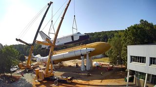 two cranes lift a full-size model of a nasa space shuttle onto its mock orange fuel tank and white solid rocket boosters in a horizontal position, with trees in the background