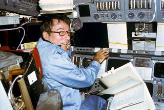 Astronaut John W. Young, mans the commander's station in the Columbia during the 36-orbit STS-1 flight. A loose leaf notebook with flight activities data floats in the weightless environment. Young is wearing a three piece constant wear flight suit. This