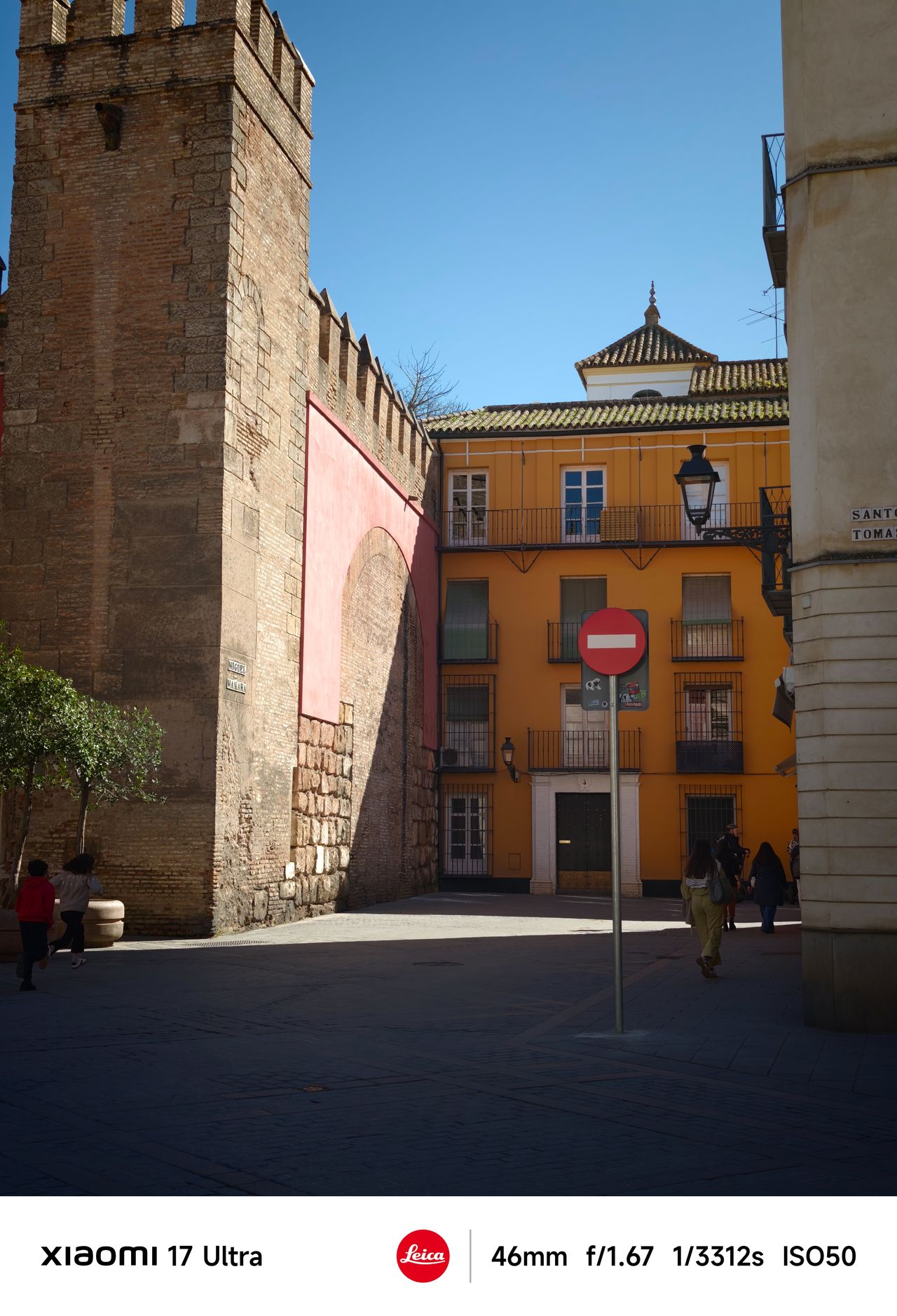 Sunlit plaza bordered by historic stone walls and a yellow building with shuttered windows, people walking across the square.