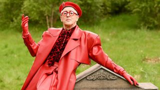 Alan Cumming is shown on The Traitors: New Blood, leaning dramatically against a gravestone, dressed in all red from his beret to his glasses, leather suit, gloves and velvet animal-print shirt.