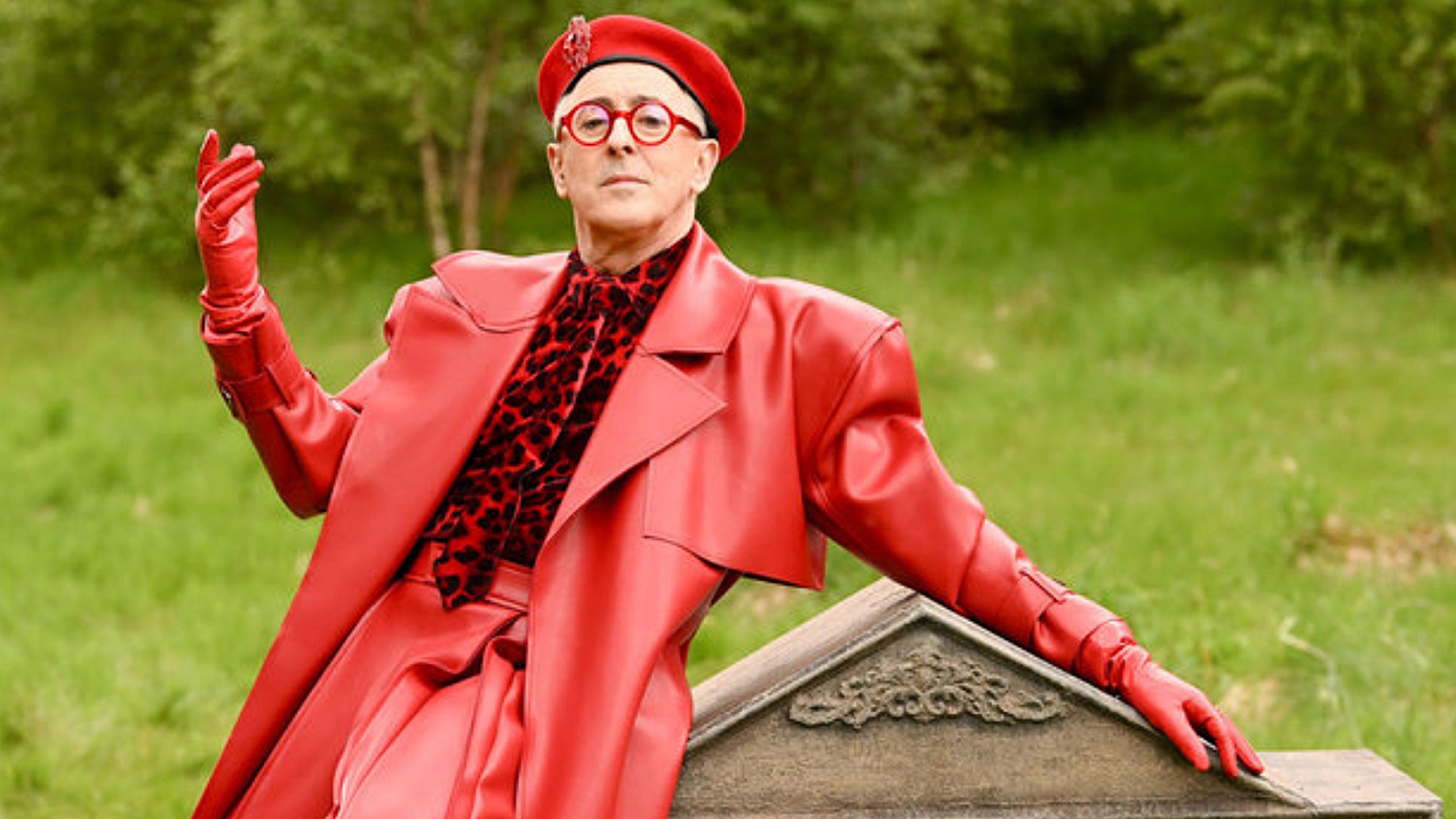 Alan Cumming is shown on The Traitors: New Blood, leaning dramatically against a gravestone, dressed in all red from his beret to his glasses, leather suit, gloves and velvet animal-print shirt.