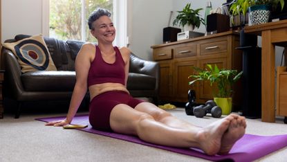 A smiling woman in sports wear sits on a yoga mat in her plant-filled living room. She is leaning back slightly, with her hands on the floor behind her, and her legs straight out in front of her.