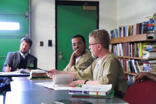 Eric Graves and another classmate sit in front of a professor around a wooden table. Both Graves and his classmate are wearing tan prison suits.